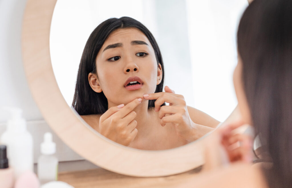Upset asian girl checking for pimples, looking at mirror