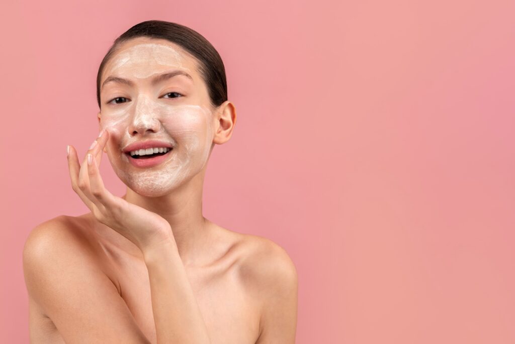 Smiling woman applying a creamy face mask as part of her skincare routine against a pink background